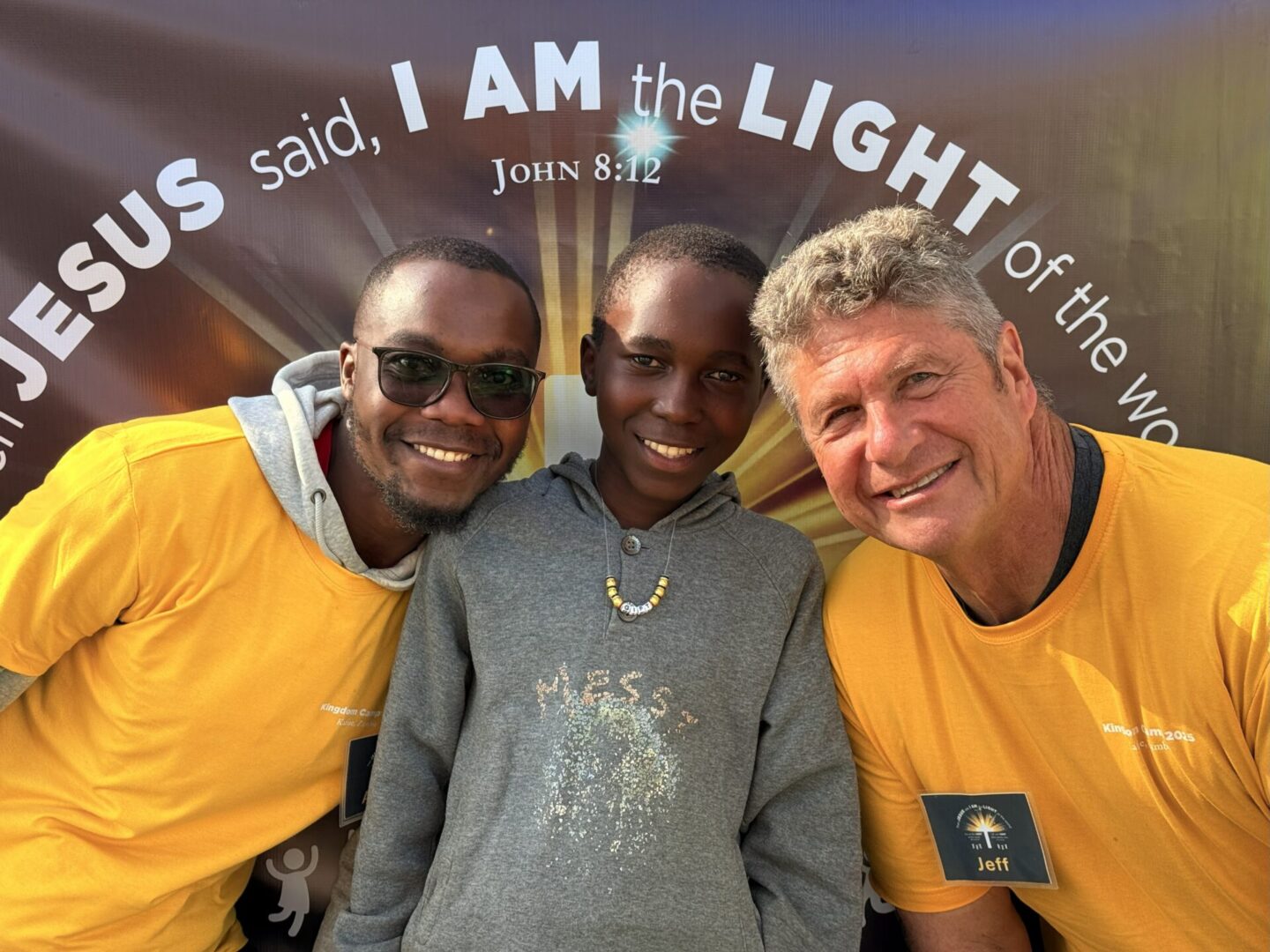 Three smiling men posing together outdoors, one wearing a bright yellow shirt.